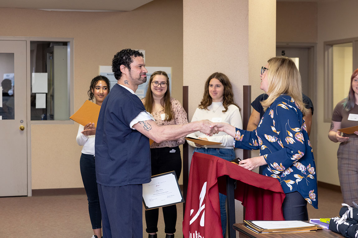 A blond woman shakes hands with a man in a navy blue prison uniform as he accepts a certificate of completion